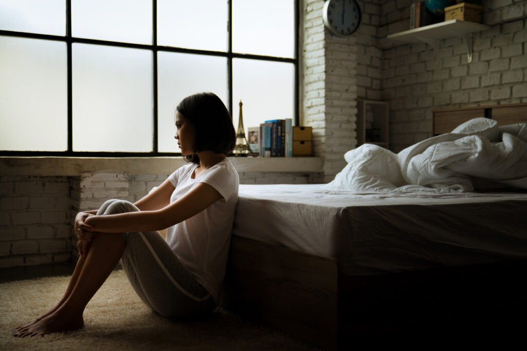 Depression and Sleep A depressed woman sits on the floor in a bedroom with her knees pulled to her chest